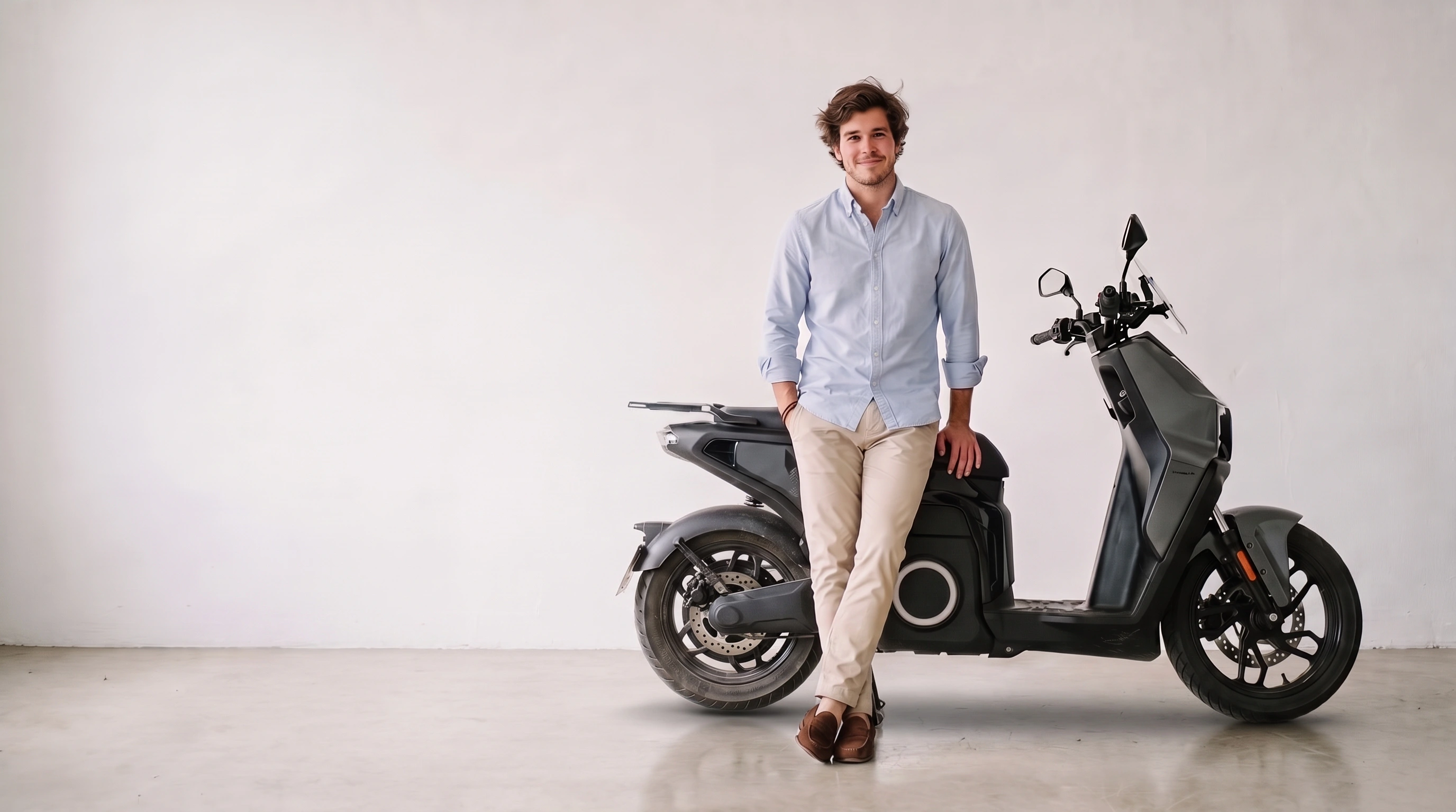 Un chico joven apoyado en una moto eléctrica Silence S02 gris, delante de una pared blanca con luz de ventana